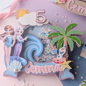May include: A layered birthday cake topper with the number 5, the name "Gemma", and characters from the movie Frozen. The design includes Anna, Elsa, Olaf, a palm tree, and a wave. The color scheme is pastel pink, blue, and white.