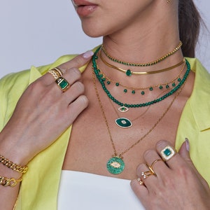 May include: A collection of gold-toned jewelry featuring emerald green accents. The pieces include necklaces, rings, earrings, and a bracelet. The necklaces have various designs, including beaded strands and pendant styles. The rings feature large emerald-cut stones.