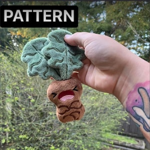 Pode incluir: Um brinquedo de pelúcia em forma de cenoura marrom com uma parte superior verde frondosa. A cenoura tem um rosto sorridente com a boca bem aberta e uma língua rosa. O brinquedo está sendo segurado em uma mão.