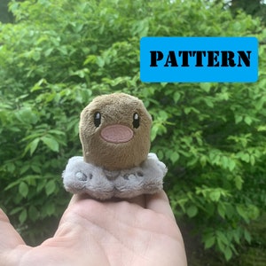 Può includere: Un giocattolo di peluche marrone e grigio di un personaggio di Pokemon. Il giocattolo ha una testa rotonda con un viso marrone e un corpo grigio. Il giocattolo è seduto su una mano.