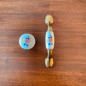 May include: A light blue ceramic cabinet knob and pull with floral designs. The knob is round, and the pull is a rectangular shape with brass hardware. The floral design includes pink, blue, and yellow flowers.