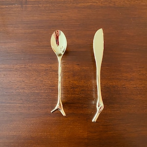 May include: A gold-colored spoon and butter knife set. The spoon has a rounded bowl and a handle that splits into three prongs at the base. The butter knife has a rounded blade and a handle that splits into three prongs at the base. The set is on a brown wooden surface.