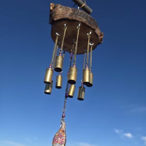 May include: A handcrafted wind chime featuring brass bullet casings suspended from a wooden base. The chime includes purple beads and a teardrop-shaped glass pendant with a floral design. The background shows a clear blue sky and a sandy landscape.