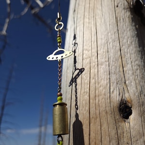 May include: A handcrafted brass earring featuring a UFO charm and a cylindrical bell. The earring hangs from a delicate chain with green beads. The background shows a weathered tree trunk and a clear blue sky.