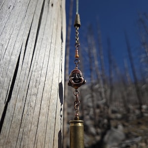 May include: A handcrafted wind chime featuring a smiling Buddha face pendant and a brass bullet casing. The chime hangs from a wooden post against a backdrop of a clear blue sky and blurred trees. The chime is made of copper-colored metal.