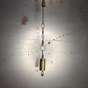 May include: A handcrafted brass bullet casing pendant with a decorative chain. The pendant features a small, cone-shaped charm, copper-colored beads, and blue beads. The pendant hangs from a silver-colored hook.