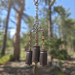 May include: A hanging ornament featuring a bronze-toned metal triquetra symbol connected to three dark brown cylindrical casings by small chains. The piece includes metallic beads and a small bronze-toned feather charm dangling from the center.