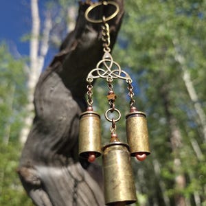 May include: A hanging ornament featuring a brass-toned metal triquetra knot connected to three cylindrical brass shell casings. The casings are suspended by small metal chains and accented with small, round, brownish-orange beads.