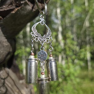 May include: A silver-toned keychain featuring a decorative filigree connector, three metallic bullet casings, and a small circular charm embossed with a compass design. The components are linked by silver chains and accented with small frosted beads.