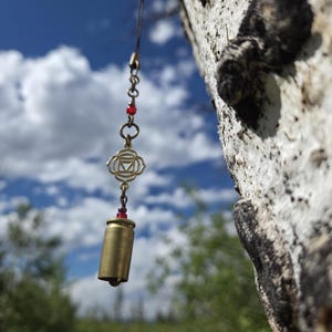 May include: A hanging ornament featuring a brass-colored bullet casing bell, a gold-toned metal charm with a triangular symbol, and small red beads. The piece is suspended by a thin dark cord from a metal hook attached to a textured tree trunk.