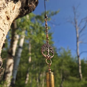 May include: A hanging ornament featuring a copper-toned tree of life charm suspended from a thin cord, connected to a metallic bullet casing at the bottom. The design includes small metallic beads along the chain, creating a vertical structure.