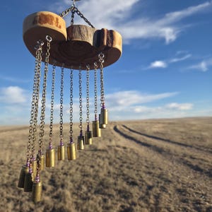 May include: A handcrafted wind chime featuring a wooden base with attached chains and brass bullet casings. The wooden base is made of natural wood slices. The chains are adorned with purple beads. The wind chime hangs from a metal ring.