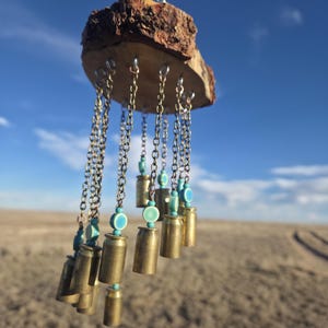 May include: A rustic wind chime featuring a natural wood base with brass bullet casings suspended by chains. Turquoise beads accent the design. The chime hangs against a backdrop of a blue sky and a field.