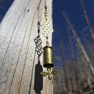 May include: A handmade brass earring featuring a honeycomb design, a bullet casing, and a small bee charm. The earring hangs from a delicate chain against a blurred background of a tree trunk and blue sky.
