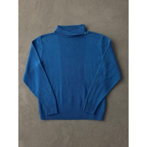 Puede incluir: Un suéter azul vibrante con un diseño de cuello único. El suéter presenta una textura acanalada en el centro y en las mangas. Un solo botón asegura el cuello. La prenda está hecha de un material suave.