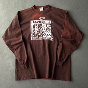 May include: A brown long-sleeve t-shirt featuring a white graphic print on the chest. The design includes the text "JAVA JOES," "COURT AVENUE DISTRICT," "DES MOINES, IOWA," and "COFFEEHOUSE EXTRAORDINAIRE" with illustrations of two figures.