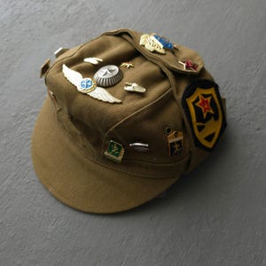 May include: A tan military-style field cap adorned with various metal pins and a fabric patch. The pins include a winged emblem, a circular badge, a tank, and small sports-themed icons. A black and yellow shield-shaped patch features a red star.