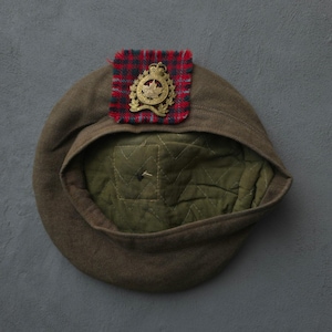 Op de afbeelding: Een bruine wollen baret met een doorgestikte olijfgroene voering. Aan de voorzijde is een vierkant lapje rood, blauw en groen ruitjesstof bevestigd, dat dient als achtergrond voor een goudkleurige metalen militaire badge met een esdoornblad.