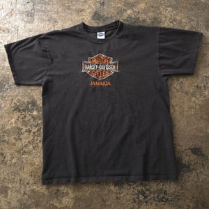 Op de afbeelding: Een zwart t-shirt met korte mouwen en het oranje-witte Harley-Davidson Motor Cycles schildlogo in het midden op de borst. Onder het logo staat het woord JAMAICA in oranje hoofdletters gedrukt. Het shirt heeft een ronde halslijn.
