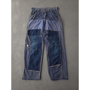 May include: Blue denim work trousers with a patchwork design. The trousers feature various shades of blue denim, with visible white paint splatters. The patchwork includes rectangular and square panels, creating a unique, distressed aesthetic. They have a button and zip closure.