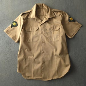 May include: A tan short-sleeve button-down shirt featuring a pointed collar, two chest pockets with buttoned flaps, and shoulder epaulets. Each sleeve displays a black patch with a yellow eagle emblem, and the front has a central button closure.