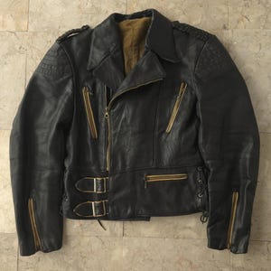 Puede incluir: Chaqueta de cuero negra con cremalleras doradas, cuello con solapas y detalles en los hombros. La chaqueta presenta cierre de cremallera diagonal, dos bolsillos delanteros y correas ajustables en la cintura con hebillas. Estilo motero clásico.