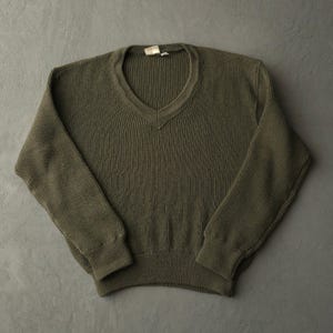 May include: An olive green long-sleeve jumper featuring a V-neckline and a ribbed knit texture throughout the body and sleeves. The garment includes a solid-coloured design with a plain hem and cuffs, showcasing a consistent knit pattern.