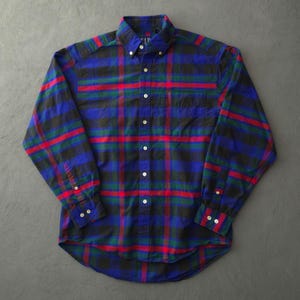 May include: A long-sleeve button-down shirt featuring a plaid pattern in blue, green, black, and red. The shirt has a button-down collar, a single chest pocket on the left side, and white buttons along the front placket and on the sleeve cuffs.