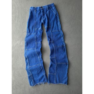 May include: A pair of blue denim jeans featuring a patchwork construction with visible vertical and horizontal seams. The pants have a button fly closure, belt loops at the waistband, and raw, unfinished frayed hems at the bottom of the legs.