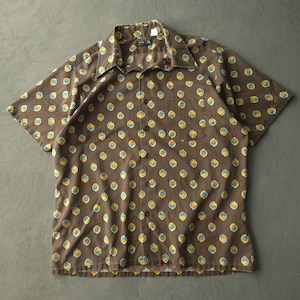 May include: A short-sleeved button-down shirt in a dark brown colour featuring a repeating pattern of small circular motifs in yellow and light blue. The shirt includes a pointed collar, a single chest pocket on the left side, and dark buttons.