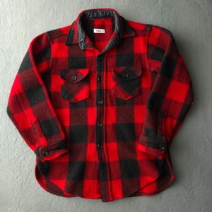 May include: A long-sleeve button-up shirt featuring a red and black buffalo plaid pattern. The shirt includes a black collar, two chest pockets with buttoned flaps, and black buttons down the front placket and on the cuffs. A white label is visible.