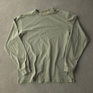 May include: A long-sleeve crew neck shirt in a muted sage green colour. The garment features a ribbed collar and a small brand label stitched inside the neckline. The fabric has a soft, slightly worn texture with visible seams along the sleeves.