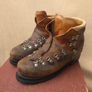 May include: A pair of brown leather hiking boots featuring a distressed finish, a high-top ankle design, silver-toned metal D-ring lace hardware, and a thick black rubber lug sole. The interior collar is lined with a light tan padded material.