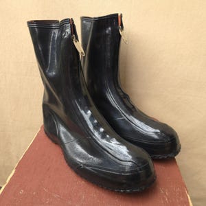 May include: A pair of black rubber overshoes featuring a high-top design with a front-facing vertical zip closure. The boots have a smooth, glossy finish, a rounded toe, and a textured sole, providing a functional and protective footwear option.