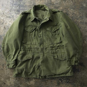 May include: An olive green field jacket featuring a fold-down collar, long sleeves with buttoned cuffs, and shoulder epaulets. The front includes a central zip closure, two chest pockets with buttoned flaps, and two lower pockets with buttoned flaps.