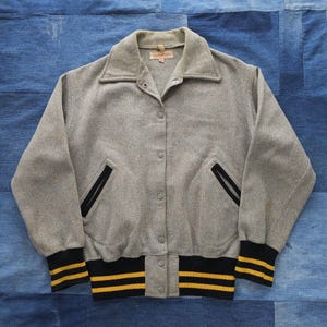 May include: A gray wool varsity jacket with black and yellow striped trim around the cuffs and waistband. The jacket has a button closure and two pockets.