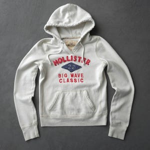 May include: A white long-sleeve hooded sweatshirt featuring a red embroidered Hollister logo, a blue diamond-shaped patch with the number 22, and the red text Big Wave Classic. The garment includes a front kangaroo pocket and white drawstrings.