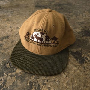 May include: A tan baseball cap with a dark olive green corduroy brim. The front features brown embroidery of a forest scene with a deer and the text "MICKEY'S OUTDOOR ADVENTURES" arched above the graphic. The cap has a structured crown.