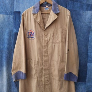 May include: A tan work jacket with blue trim on the collar and cuffs. The jacket has a large patch on the left chest that says "GM Service".