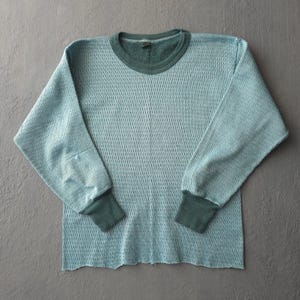 May include: A long-sleeve thermal shirt in a light blue colour with a textured waffle-knit pattern. The garment features a dark green ribbed crew neckline and matching dark green ribbed cuffs at the end of each sleeve, providing a contrast.