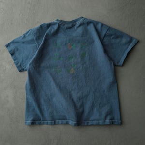 Op de afbeelding: Een slate blauw t-shirt met korte mouwen en een raster van kleine, gedempte botanische illustraties op de achterkant. Het kledingstuk heeft een ronde hals en zichtbare witte stiksels langs de kraag en zoom voor een subtiel contrast.