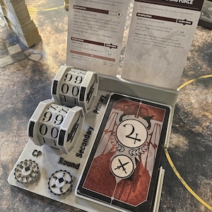 May include: A tabletop game setup featuring cards, dice, and counters. The cards have text and graphics, while the dice display numbers. The counters show numbers and symbols. The game appears to be a strategy or war game.