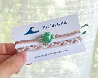 May include: Two handmade bracelets are displayed on a card that reads "BLUE TIDE CRAFTS". One bracelet has a green turtle charm, and the other is a braided design. The card also has a wave graphic.