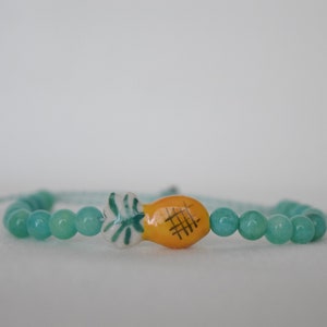 May include: A turquoise beaded bracelet with a central ceramic pineapple charm. The pineapple is yellow with black crosshatch detailing. The bracelet is strung on a light blue cord. The beads are approximately 0.6 cm in diameter.