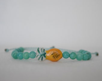 May include: A beaded bracelet featuring turquoise-colored beads and a central ceramic charm shaped like a yellow pineapple with black crosshatch detailing. The bracelet is strung on a light blue cord.