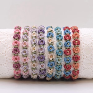May include: A collection of daisy chain bracelets in various colors, including pink, purple, blue, and orange. Each bracelet features small, crocheted flower details and a green cord. The bracelets are displayed on a white, textured surface.