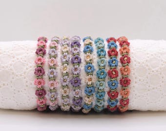 May include: A collection of daisy chain bracelets in various colors, including pink, purple, blue, and orange. Each bracelet features small, crocheted flower details and a green cord. The bracelets are displayed on a white, textured surface.