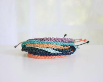May include: A stack of braided rope bracelets in various colors, including purple, teal, orange, and navy. The bracelets are handmade and appear to be made of a woven material. The bracelets are stacked on top of each other.