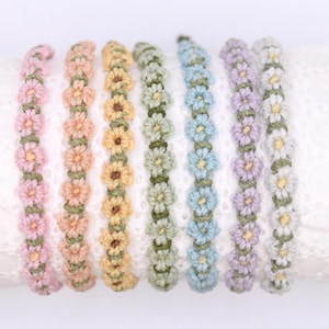 May include: A selection of daisy chain bracelets in pastel shades. Each bracelet is adorned with crocheted flowers in pink, peach, yellow, green, blue, purple, and white, with green stems. The bracelets are displayed on a white lace surface. The bracelets are handmade.