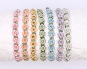 May include: A collection of handmade daisy chain bracelets in various pastel colors. Each bracelet features a row of crocheted flowers in shades of pink, peach, yellow, green, blue, purple, and white, with green stems. The bracelets are displayed on a white lace surface.